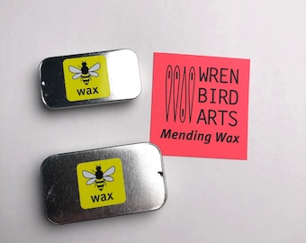 May include: Two silver tins with yellow labels and a black bee illustration. The labels read "wax". A red square with white text reads "WREN BIRD ARTS Mending Wax".