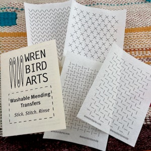 May include: Four white sheets of washable mending transfers with different patterns: chevron, crosshatch, and two different grid patterns. The sheets are stacked on top of a book with the text "WREN BIRD ARTS Washable Mending Transfers Stick. Stitch. Rinse" and the website address "wrenbirdarts.com".