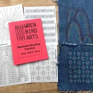 Op de afbeelding: Een roze pamflet met de titel "WREN BIRD ARTS" en de tekst "Washable Mending Transfers Stick. Stitch. Rinse". Het pamflet ligt op een houten oppervlak met verschillende vellen papier met verschillende stikpatronen.