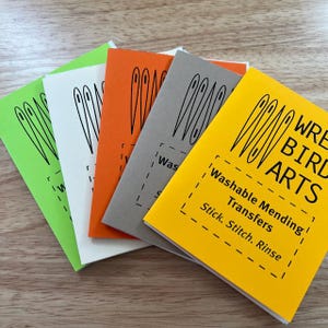 Puede incluir: Cinco cuadernos de papel de colores vivos con ilustraciones de agujas negras y el texto "WRE BIRD ARTS" en el cuaderno amarillo. El cuaderno amarillo también tiene el texto "Transferencias de costura lavables Pegar. Coser. Enjuagar".