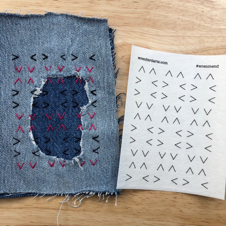 May include: A denim patch with a hole in the centre, surrounded by black and pink stitching in a chevron pattern. The patch is next to a sheet of paper with the text "wrenbirdarts.com" and "#wrenmend" printed on it. The paper is covered in black chevron patterns.