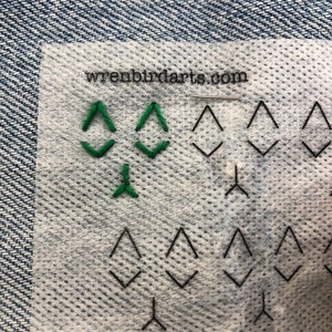 May include: A close-up of a white fabric with a green embroidered design of three triangles. The fabric has a grid pattern and the text "wrenbirdarts.com" is printed in black at the top.