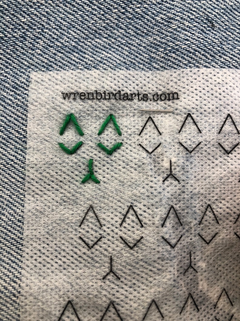 May include: A close-up of a white fabric with a green embroidered design of three triangles. The fabric has a grid pattern and the text "wrenbirdarts.com" is printed in black at the top.