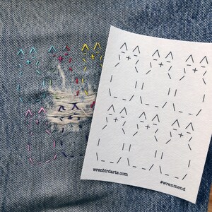 May include: A close-up of denim fabric with a hole repaired with colourful embroidery. A white piece of paper with a black and white pattern is placed on top of the denim. The paper has the text "wrenbirdarts.com" and "#wrenmend" printed on it.