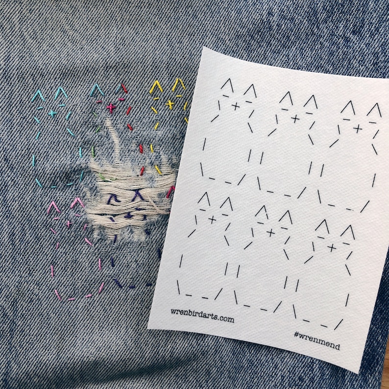 May include: A close-up of denim fabric with a hole repaired with colourful embroidery. A white piece of paper with a black and white pattern is placed on top of the denim. The paper has the text "wrenbirdarts.com" and "#wrenmend" printed on it.