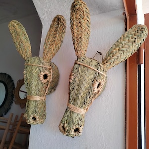 Puede incluir: Dos adornos de pared con forma de cabeza de burro tejidos. Las cabezas de burro están hechas de paja tejida en tonos verdes y beige. Tienen orejas largas y un agujero para el ojo. Las cabezas de burro están sujetas a la pared con cuerda.