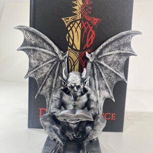 Può includere: Una statuetta di gargoyle color argento con grandi ali dettagliate, accovacciata mentre legge un libro aperto. La figura è posizionata davanti a un libro con copertina rigida nera con un grafico di una spada oro e rosso e il testo rosso "D...CE".