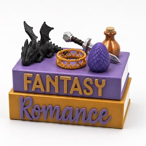 Può includere: Una pila di due libri con le parole FANTASY e Romance sui dorsi. Il libro viola superiore presenta una statuetta di drago nero, un anello dorato con motivo a cuori, una spada argentata, un uovo viola testurizzato e una piccola bottiglia color rame.