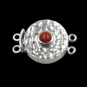 May include: A silver-colored jewelry clasp with a hammered texture and a central red bead. The clasp has a round shape with two loops on each side for attachment. The background is black.