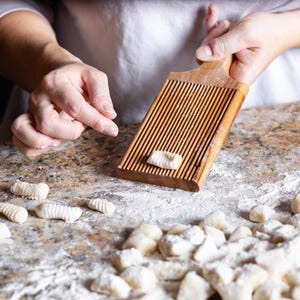 May include: A wooden gnocchi board with parallel grooves is held by a person, with a single gnocchi resting on it. Several uncooked gnocchi are scattered on a floured surface, ready for cooking. The scene suggests homemade pasta preparation.