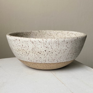 May include: A white ceramic bowl with brown speckles and a beige rim. The bowl is sitting on a white surface.