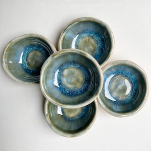 Op de afbeelding: Een verzameling van vijf handgemaakte keramische kommen. Elke kom heeft een unieke mix van blauw, groen en bruin glazuur, wat een gemarmerd effect creëert. De kommen hebben een afgeronde vorm met een licht verhoogde rand, perfect voor dips of kleine porties.