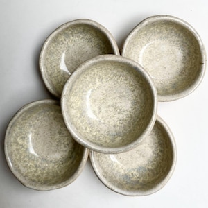 Pode incluir: Cinco pequenas tigelas redondas de cerâmica com um esmalte salpicado branco-sujo e verde-claro. As tigelas estão dispostas em um padrão circular, mostrando seu design simples. Perfeitas para servir molhos ou pequenas porções.