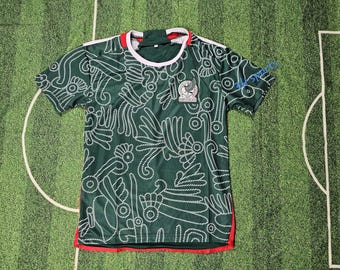 May include: Green soccer jersey with white and red accents. The jersey features a white pattern and the Mexican national team emblem. The collar is white with a red trim, and the sleeves have white stripes.