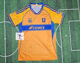 May include: A yellow and blue soccer jersey with a V-neck collar. The jersey features blue stripes on the shoulders and a blue stripe across the chest with the word "CEMEX". Logos and text are visible on the front.