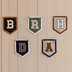 May include: Five felt pennant banners, each displaying a different letter: B, R, H, D, and A. The banners feature various color combinations, including black and gold, green and white, brown and white, navy and brown, and navy and tan. Each banner is hung with metal grommets.