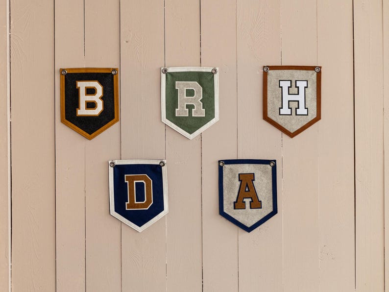 May include: Five felt pennant banners, each displaying a different letter: B, R, H, D, and A. The banners feature various color combinations, including black and gold, green and white, brown and white, navy and brown, and navy and tan. Each banner is hung with metal grommets.