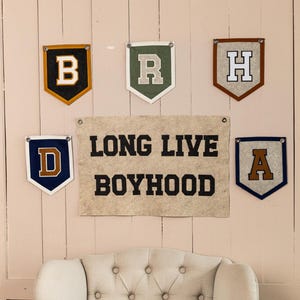 May include: Wall decor featuring felt pennants with letters B, R, H, D, and A, along with a banner that reads "LONG LIVE BOYHOOD." The pennants have various color schemes, including gold, green, brown, and blue.