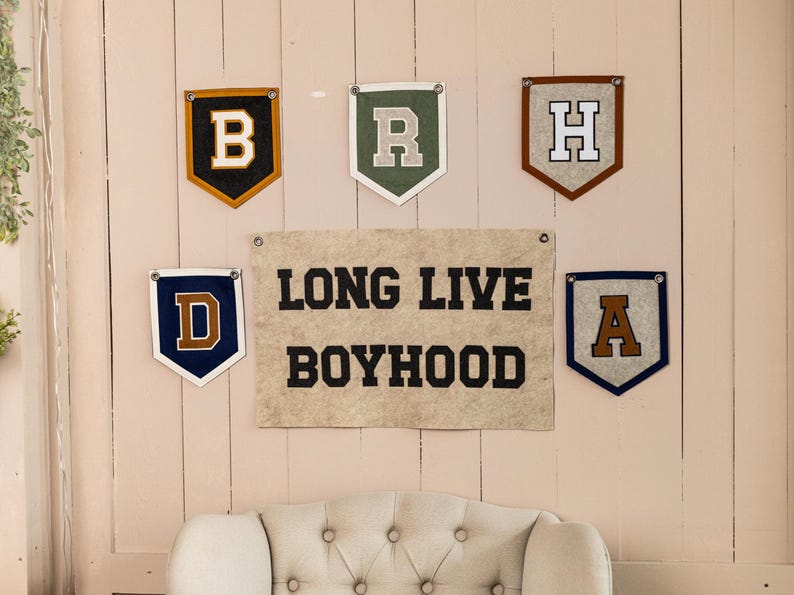 May include: Wall decor featuring felt pennants with letters B, R, H, D, and A, along with a banner that reads "LONG LIVE BOYHOOD." The pennants have various color schemes, including gold, green, brown, and blue.