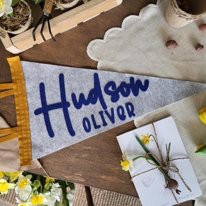 May include: A gray felt pennant with the name "Hudson Oliver" in navy blue script. The pennant has a gold trim and is displayed with a floral arrangement and a card with a daffodil.