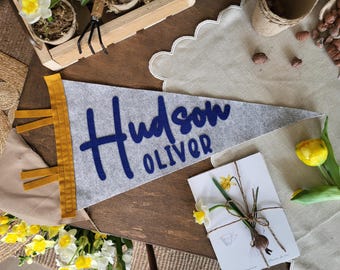 May include: A triangular gray felt pennant featuring the name Hudson Oliver in dark blue felt lettering. The pennant has a mustard yellow vertical strip on the left side with three matching yellow fabric ties extending from the edge of the strip.