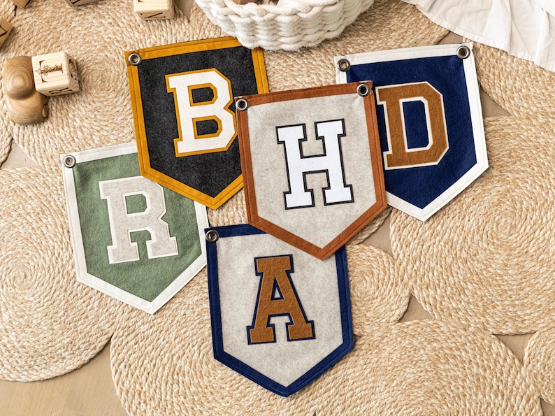 May include: Five felt pennant banners with letters B, H, D, R, and A. The banners are in various colors, including black, navy, green, and tan, with contrasting letter colors. Each banner has a metal grommet for hanging. They are arranged on a woven mat.