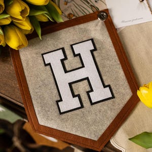 May include: A felt pennant with a brown border and a large white letter "H" with a black outline. The pennant is light gray and has a button at the top. Yellow tulips and a single yellow tulip are in the background.