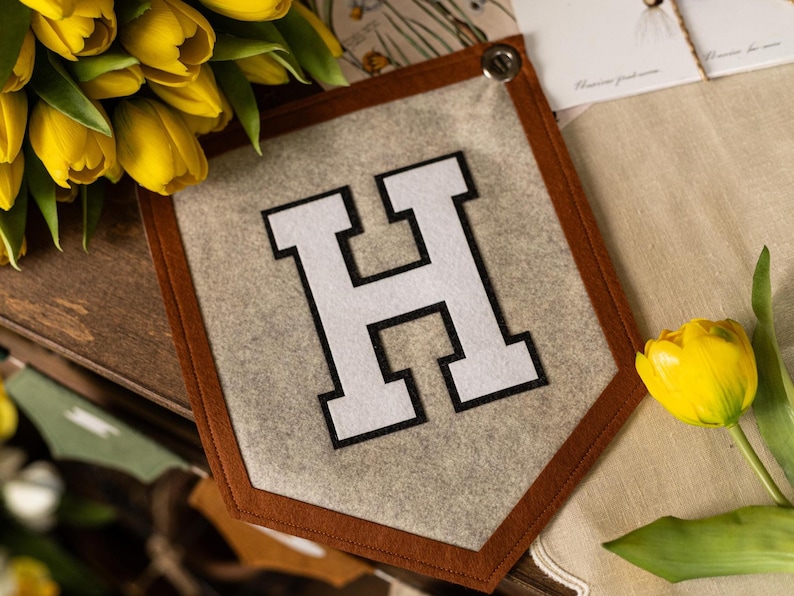 May include: A felt pennant with a brown border and the letter "H" in white with a black outline. The pennant is gray and has a button at the top. Yellow tulips are in the background.
