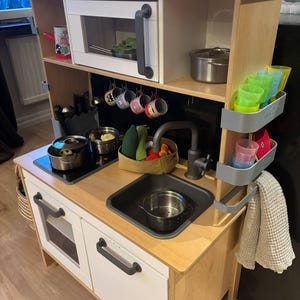 Könnte beinhalten: Eine Spielzeugküche aus Holz mit weißer Mikrowelle, Backofen und Spüle. Sie enthält ein Kochfeld, Töpfe, Pfannen und farbenfrohes Zubehör wie Tassen und ein Geschirrtuch. Die Küche ist für Kinderspiele konzipiert.