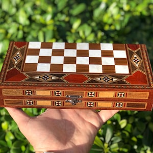 May include: A wooden chess set box with a checkerboard top. The box is made of various wood tones and inlaid with decorative patterns. The chess board squares alternate between brown and white. A metal clasp secures the box.