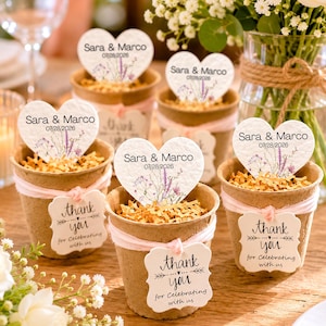 May include: Five small brown pots filled with shredded paper, each featuring a heart-shaped card printed with 'Sara & Marco 07/28/2026' and a floral design, plus a square tag tied with pink string that reads 'thank you for celebrating with us'.