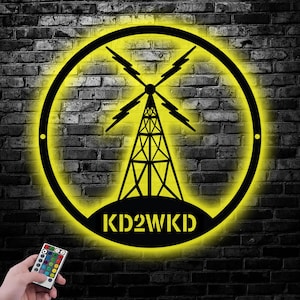 Peut inclure: Panneau lumineux noir et jaune représentant une tour radio dans un cercle. Le texte "KD2WKD" est inscrit en bas. Le panneau est fixé sur un mur de briques. Une télécommande avec des options de couleurs est au premier plan.