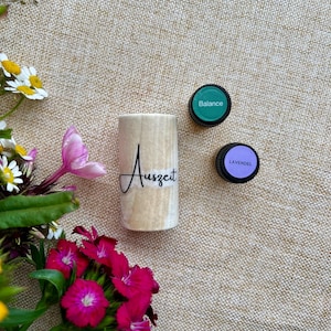 Könnte beinhalten: Eine hellfarbene Holzflasche für ätherische Öle mit dem Wort "Auszeit" in Schriftform. Zwei kleine, runde Flaschen mit schwarzen Deckeln sind mit "Balance" und "Lavendel" beschriftet. Das Bild enthält rosa und weiße Blumen.