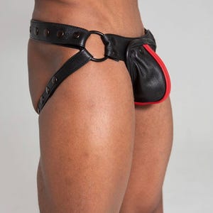 Puede incluir: Jockstrap de cuero negro con ribete rojo. El jockstrap presenta una cinturilla ancha con detalles de remaches, un anillo circular y correas que se aseguran alrededor de los muslos. La bolsa tiene una franja roja a lo largo del borde.