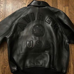 Könnte beinhalten: Schwarze Lederjacke mit dem Wort "AVIREX" auf dem Rücken, über einem kreisförmigen Emblem und dem Wort "USA". Die Jacke hat einen gerippten Saum und Bündchen sowie einen Kragen.