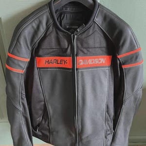 Op de afbeelding: Zwarte leren motorjas met rode strepen en het "HARLEY-DAVIDSON" logo over de borst. De jas heeft een volledige ritssluiting en hangt aan een zwarte hanger.