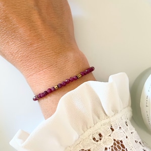 May include: A delicate bracelet featuring small, faceted, deep red gemstones strung together. The bracelet includes small gold-colored spacers. The bracelet is worn on a wrist, against a backdrop of a white, lacy sleeve.
