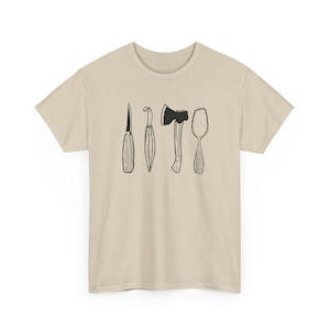 May include: A beige short-sleeve t-shirt featuring a black line art graphic on the chest. The illustration displays four distinct woodworking tools arranged in a row: a straight carving knife, a hook knife, a small hatchet, and a wooden spoon.