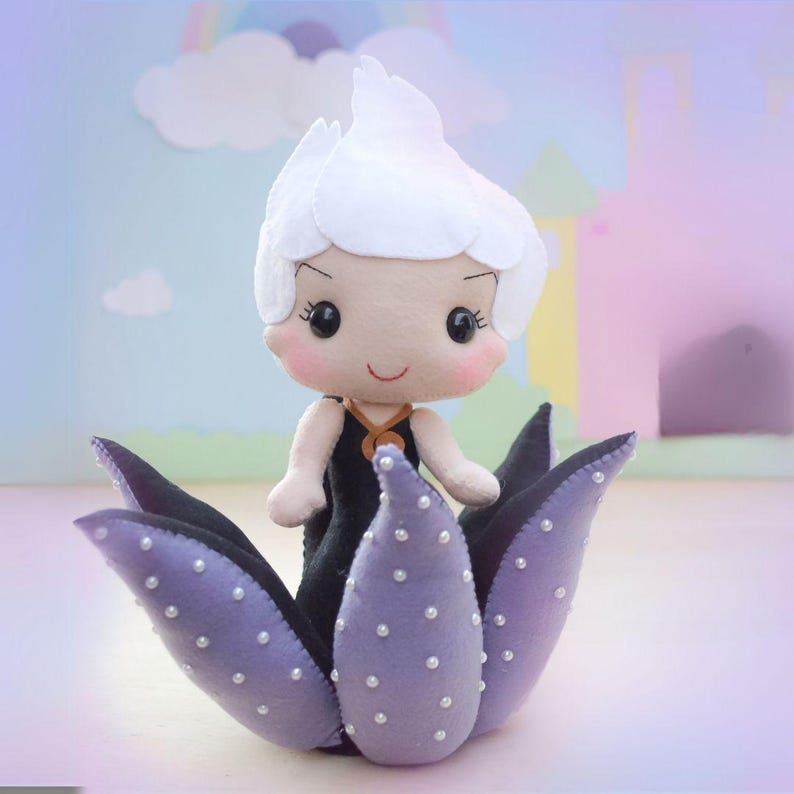May include: A handmade felt doll of a character with white hair, wearing a black dress, and standing in a purple flower. The doll has rosy cheeks and a smiling expression. The flower petals are adorned with small white beads.
