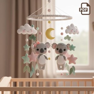 May include: A nursery mobile with two grey koala bears, clouds, stars, and a yellow crescent moon. The mobile hangs from a white ring and has pink, green, and yellow details. The koalas have pink bows and are surrounded by felt shapes.