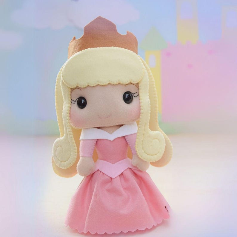 May include: A handmade felt doll of a princess, featuring a pink dress, blonde hair, and a brown crown. The doll has a sweet expression with large black eyes and is set against a soft, pastel background.