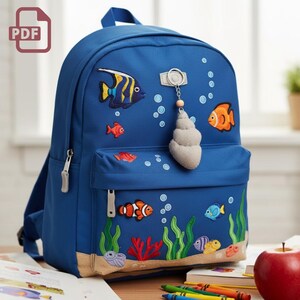 May include: Blue backpack with embroidered aquatic-themed designs, including colorful fish, seaweed, and bubbles. Features a front pocket, a seashell charm, and a PDF icon in the upper left corner. Zippers and straps are visible.