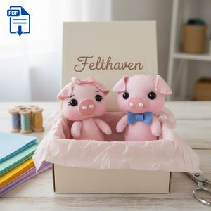 May include: Two pink felt pig stuffed animals with black eyes and pearl accents sit in a box lined with pink tissue paper. One pig has a bow, the other a blue bow tie. The box has the word "Felthaven" on it.