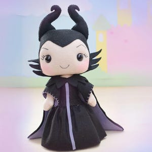 May include: A handmade plush doll of a character with black horns, a black dress, and a purple-lined cape. The doll has a smiling face with rosy cheeks and black eyes. The background is a soft pastel scene with a castle.
