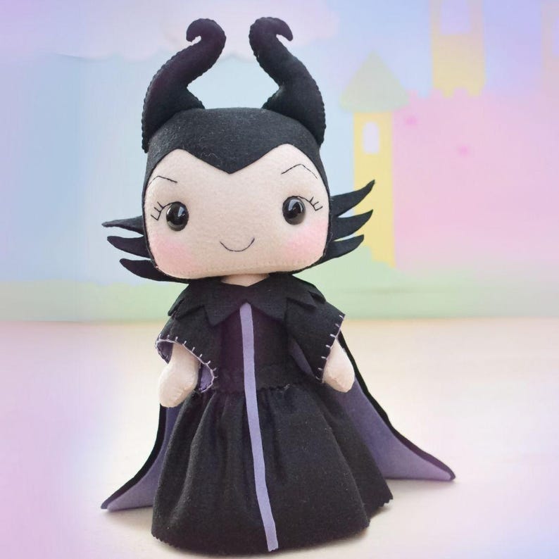 May include: A handmade plush doll of a character with black horns, a black dress, and a purple-lined cape. The doll has a smiling face with rosy cheeks and black eyes. The background is a soft pastel scene with a castle.