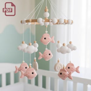 May include: A baby mobile featuring pink felt fish, white clouds, and wooden beads. The mobile is suspended from a wooden ring and hangs above a white crib. The design is soft and whimsical.