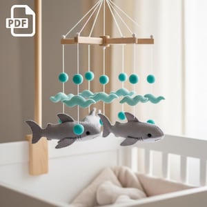 May include: A baby mobile featuring gray felt sharks, teal clouds, and teal pom-poms suspended from a wooden frame. The mobile is designed to hang above a crib or bed. The PDF icon is in the top left corner.