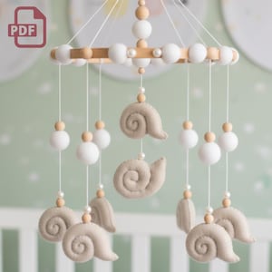 Op de afbeelding: Een baby mobile met een houten frame, witte vilten ballen en beige vilten slakvormen. De mobile wordt opgehangen met witte touwtjes en bevat houten kralen en parelaccenten. Het ontwerp is zacht en neutraal, geschikt voor een kinderkamer.