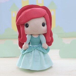May include: A handmade doll of a princess with red hair, wearing a teal dress. The doll has a light skin tone, large black eyes, and a sweet expression. The doll's hair is styled in a classic princess look. The background features a castle and clouds.