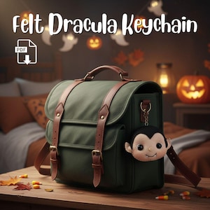May include: A green satchel with brown leather straps and a felt Dracula keyring. The keyring features a smiling vampire face. The text "Felt Dracula Keychain" is at the top of the image.